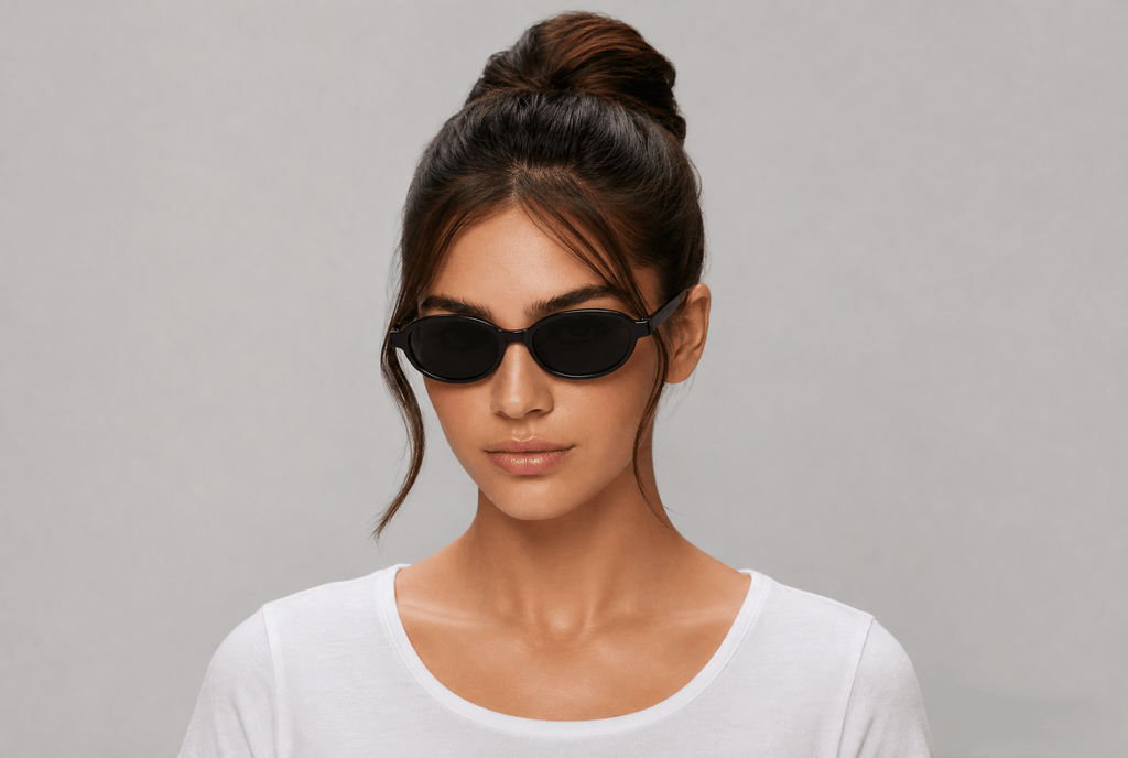 A woman with her hair in a high bun wearing black cat-eye sunglasses and a white top, photographed against a neutral gray background.