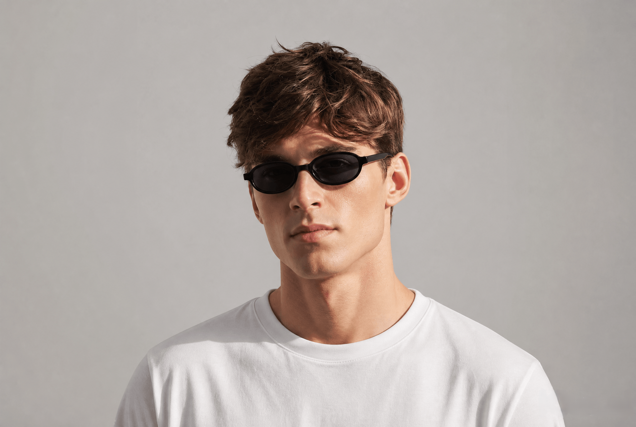 A man with short brown hair wearing black oval sunglasses and a plain white t-shirt, photographed against a neutral light gray background.