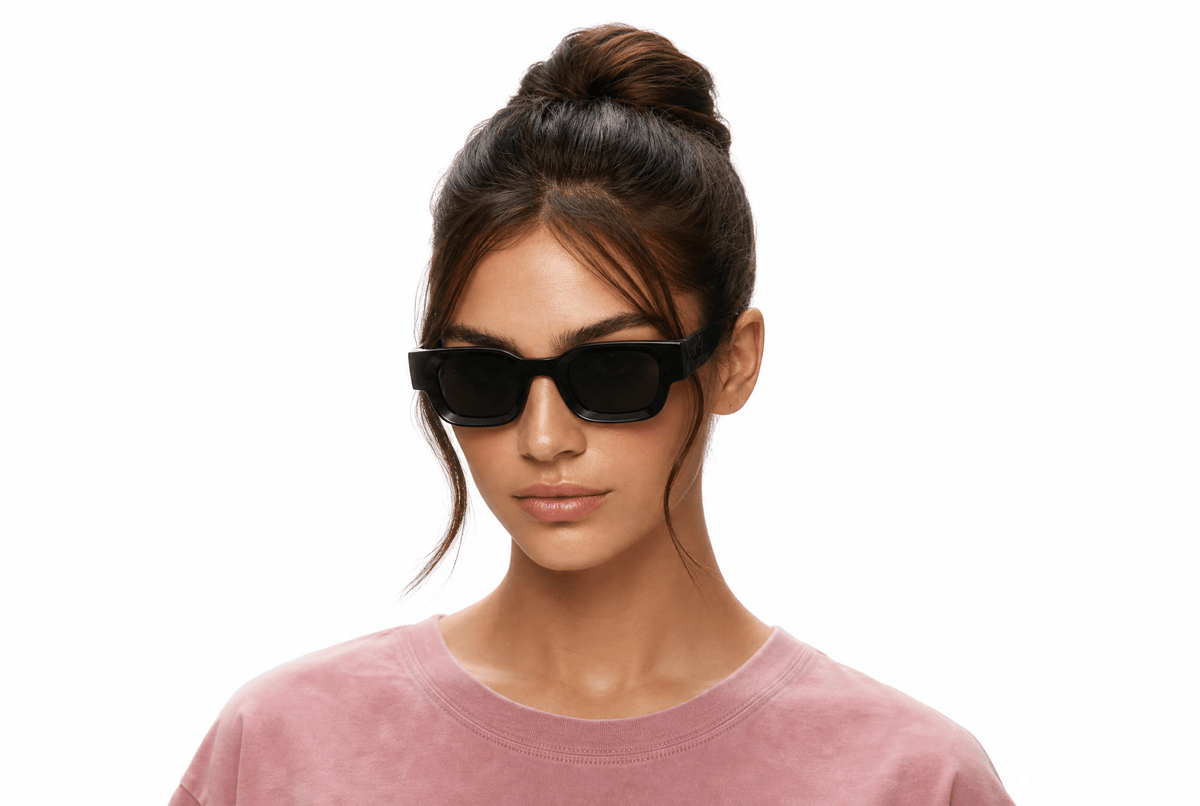 A woman with her hair in a high bun wearing black rectangular sunglasses and a dusty rose crewneck sweatshirt, photographed against a solid white background. Black