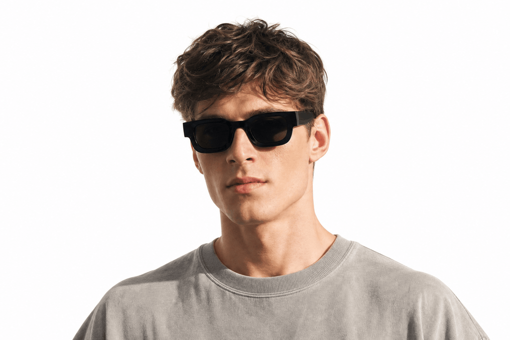 A young man with wavy brown hair wearing black rectangular sunglasses and a light gray crewneck t-shirt, photographed against a solid white background. Balck