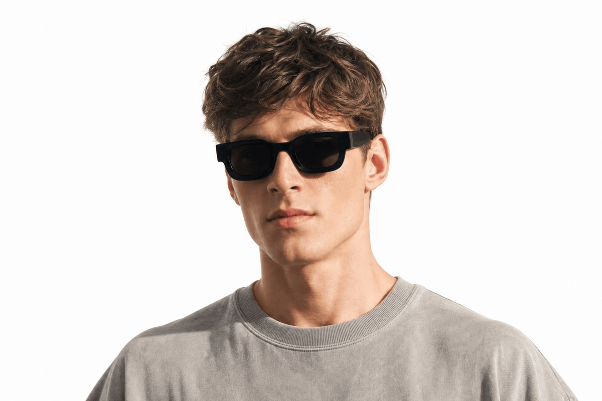 A young man with wavy brown hair wearing black rectangular sunglasses and a light gray crewneck t-shirt, photographed against a solid white background. Balck