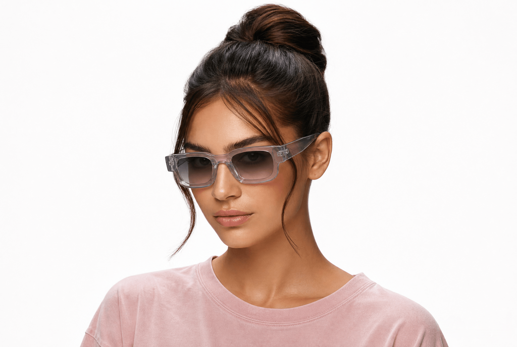 A woman with her hair in a high bun wearing translucent gray square sunglasses and a light pink crewneck shirt, photographed against a solid white background. Clear