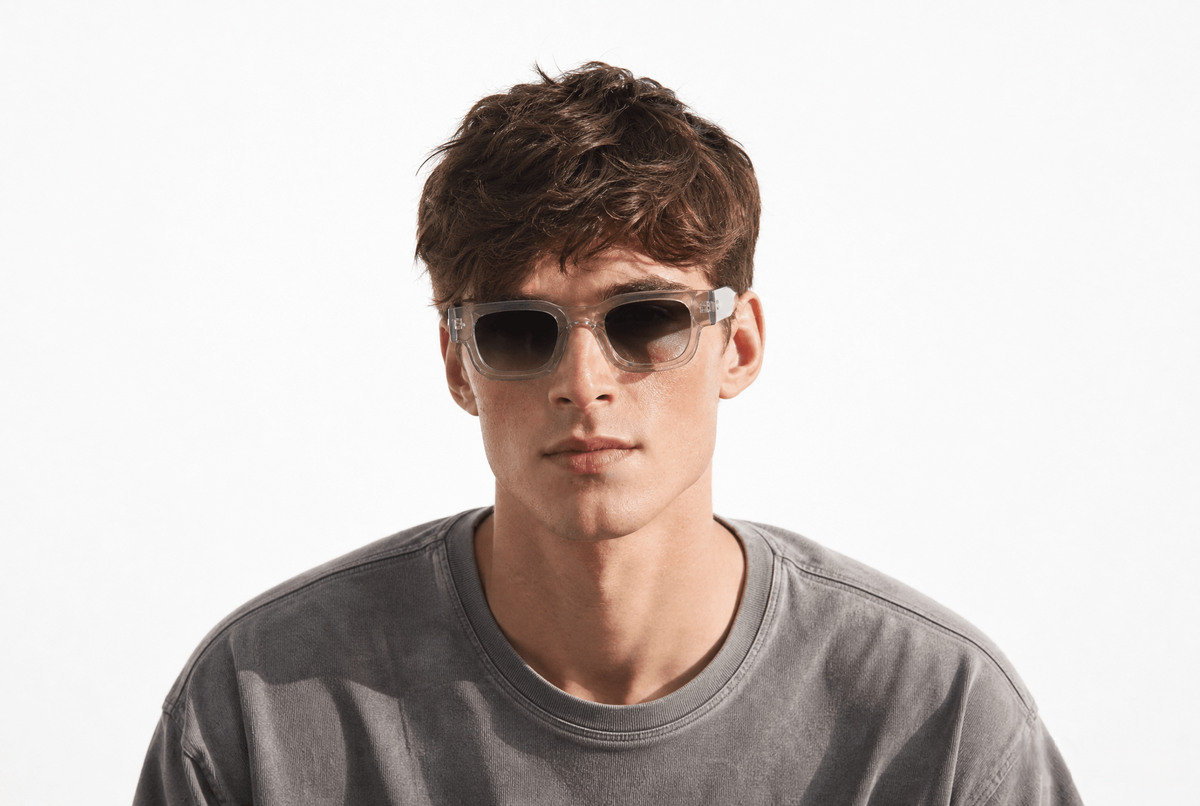 A young man with wavy brown hair wearing clear-framed square sunglasses with dark lenses and a gray crewneck t-shirt, photographed against a clean white background. Clear
