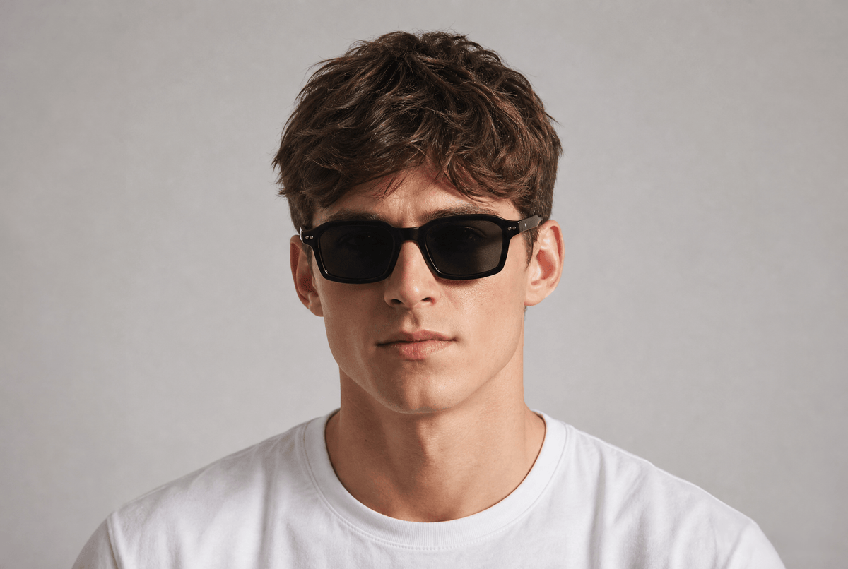 A man with wavy brown hair wearing black rectangular sunglasses and a white crew-neck t-shirt, photographed against a neutral light gray background. Black