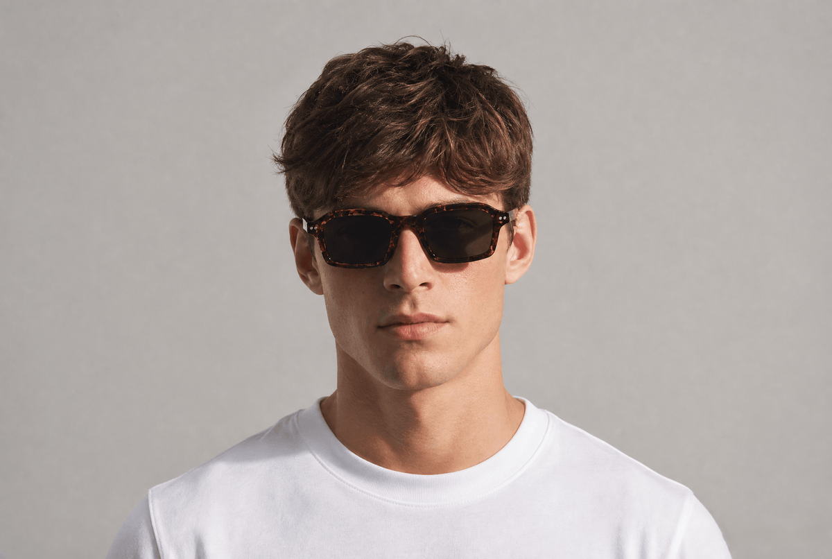 A man wearing dark tortoiseshell sunglasses and a white t-shirt, photographed against a neutral light gray background. Tortoiseshell Leo