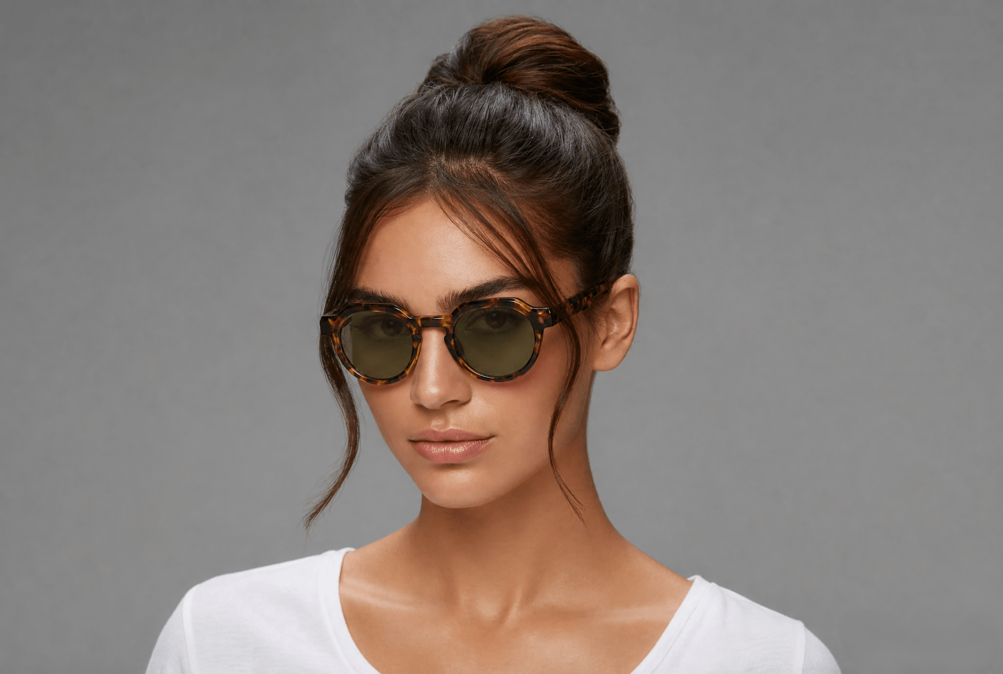 A woman with her hair in a high bun wearing round sunglasses with tortoiseshell frames and dark lenses, set against a solid gray background.