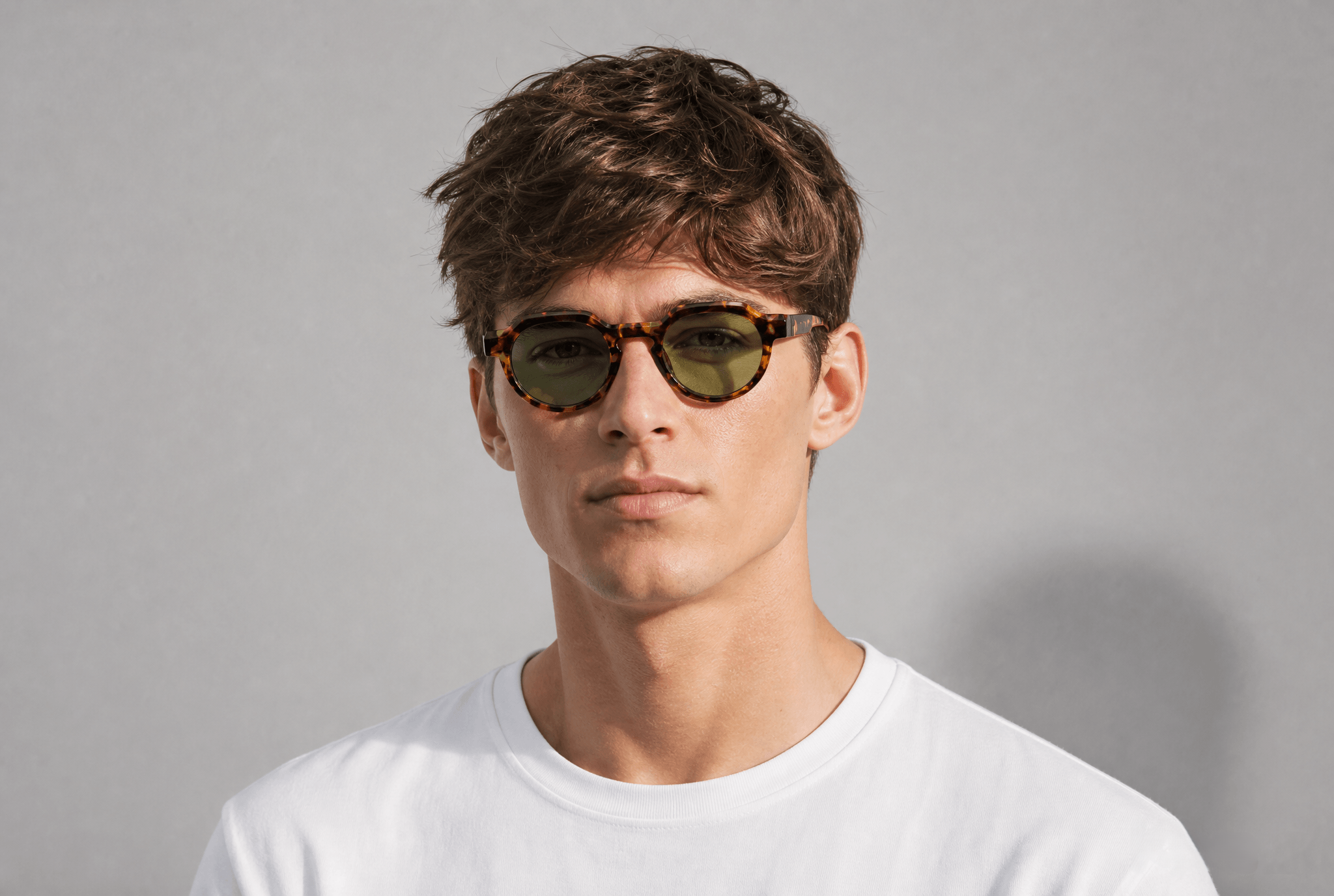 A man wearing tortoiseshell sunglasses with olive green lenses and a white crewneck t-shirt, photographed against a neutral gray background.