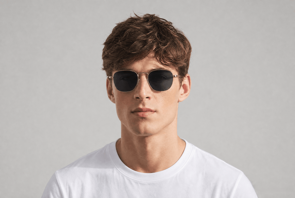 A man wearing round black sunglasses with thin gold metal frames and a white t-shirt, photographed against a neutral light gray background.