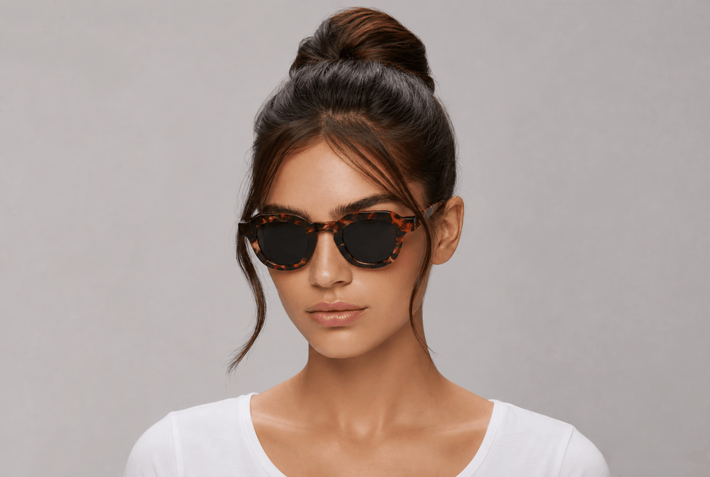 A woman with her hair in a high bun wearing tortoiseshell sunglasses with dark lenses, set against a plain gray studio background.