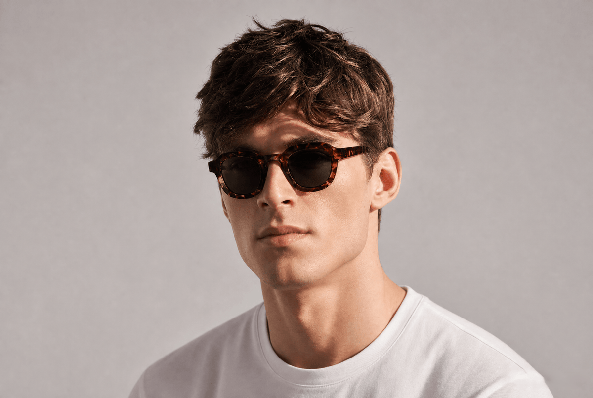 A man wearing round tortoiseshell sunglasses and a white t-shirt, photographed against a neutral light gray background.