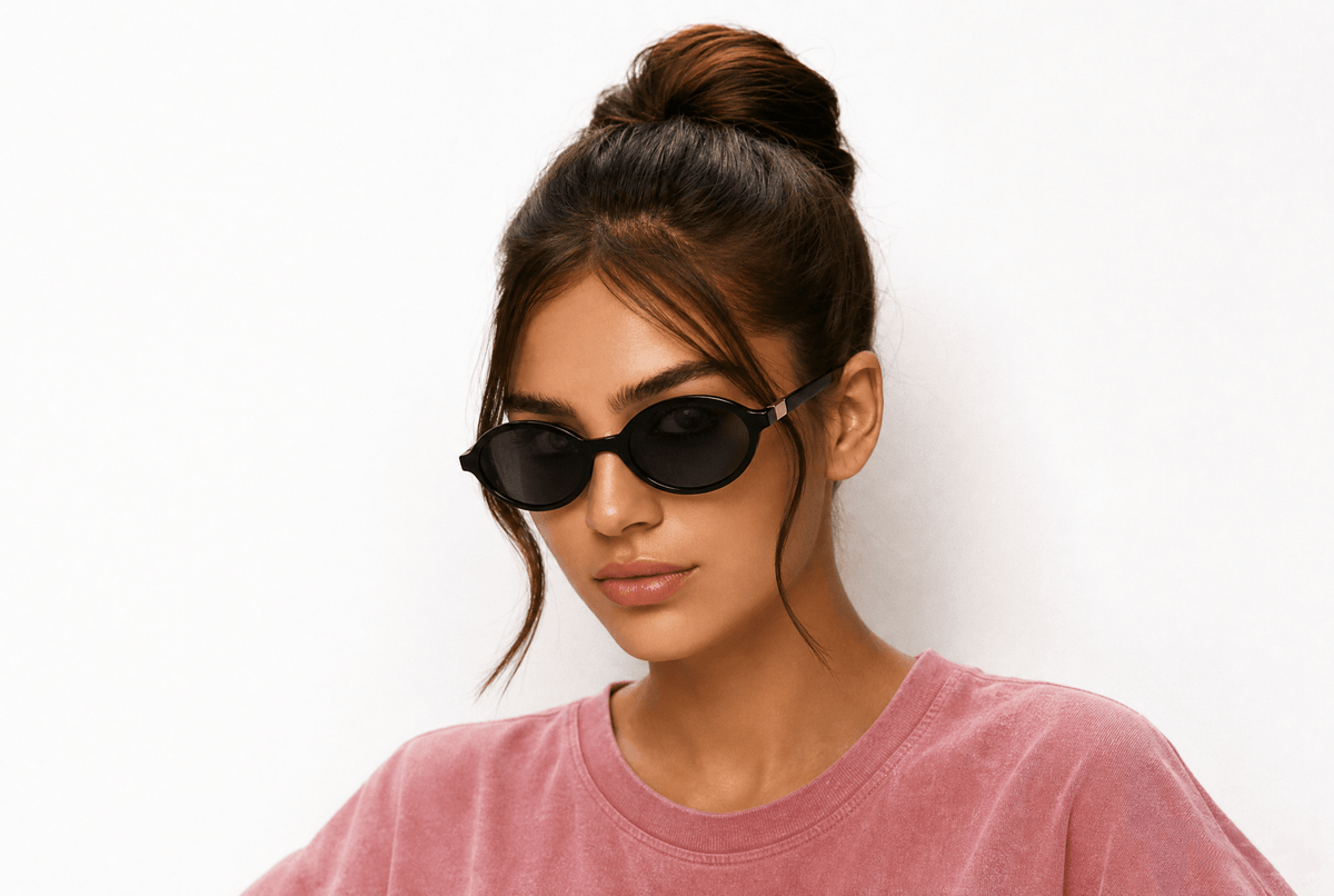 A young women wearing oval black sunglasses and a pink crewneck t-shirt, photographed against a clean white studio background. Black