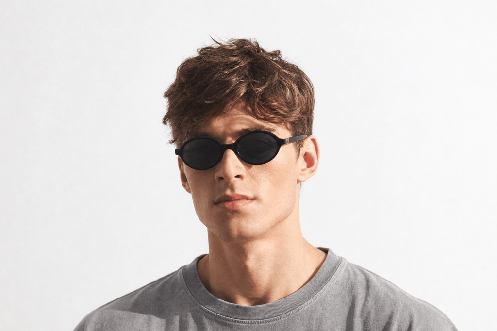 A young man with short brown hair wearing oval sunglasses and a gray crewneck t-shirt, photographed against a clean white studio background. Black