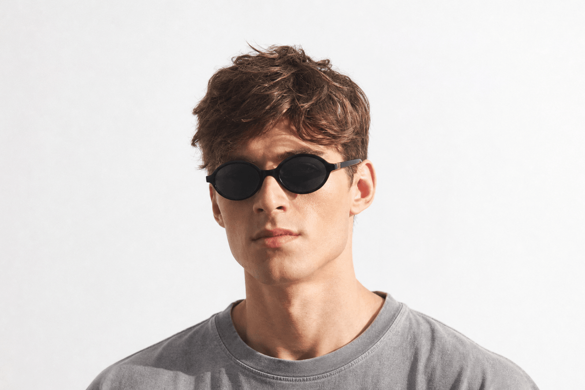 A young man with short brown hair wearing oval sunglasses and a gray crewneck t-shirt, photographed against a clean white studio background. Black