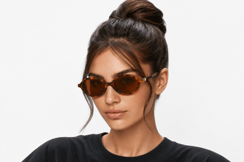 A young women wearing oval sunglasses and a dark crewneck t-shirt, photographed against a clean white studio background. Tortoiseshell