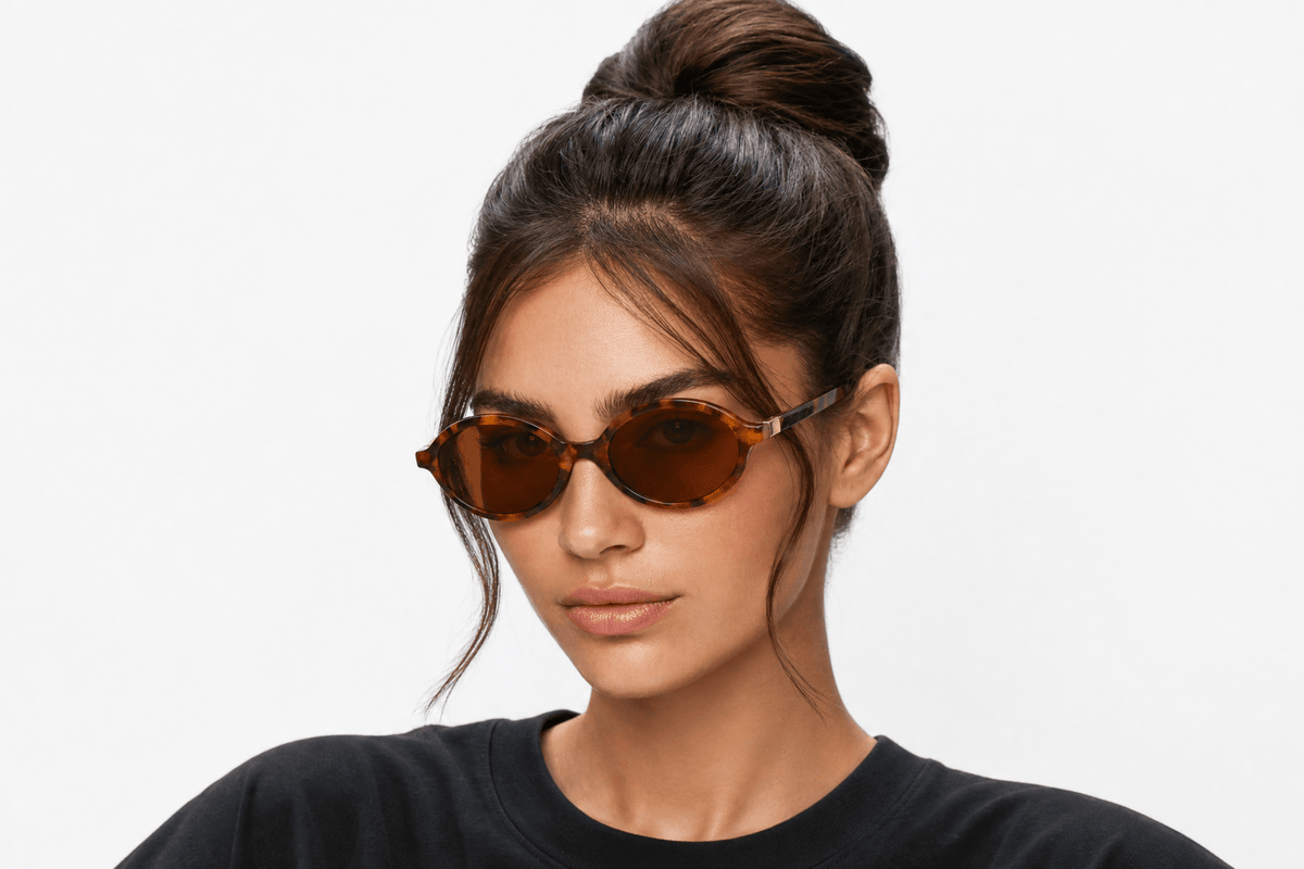 A young women wearing oval sunglasses and a dark crewneck t-shirt, photographed against a clean white studio background. Tortoiseshell