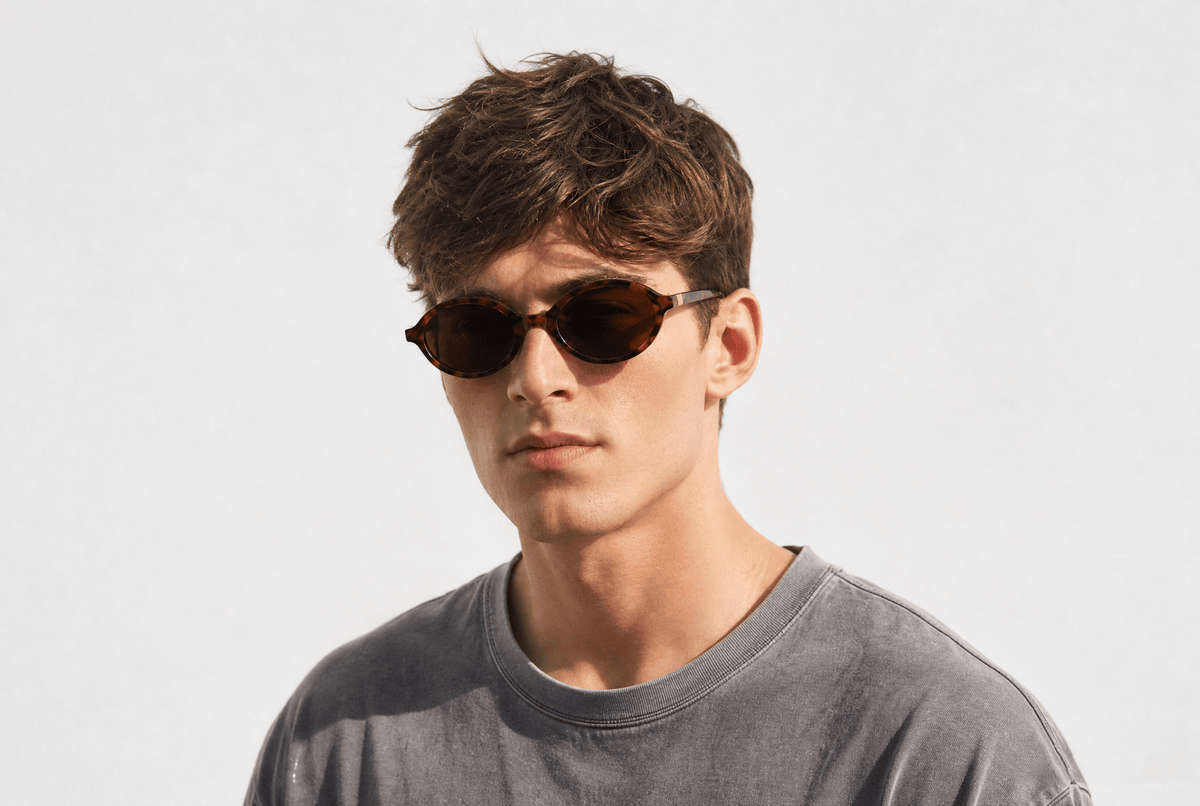A young man with short brown hair wearing oval sunglasses and a gray crewneck t-shirt, photographed against a clean white studio background. Tortoiseshell
