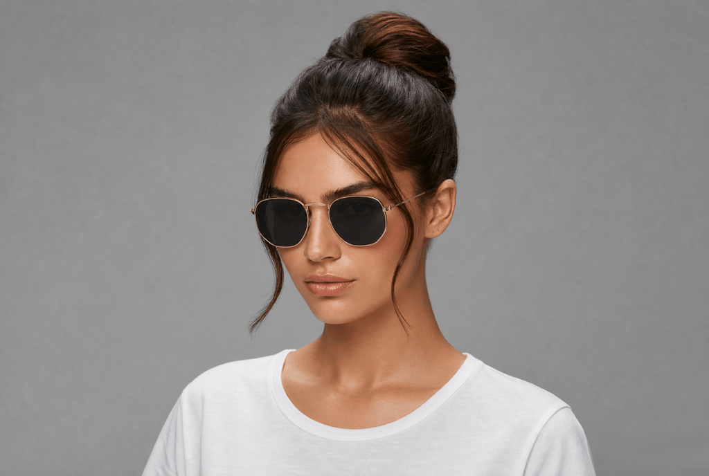 A woman with her hair in a high bun wearing round metal-framed sunglasses with dark lenses and a white t-shirt, set against a solid gray background.