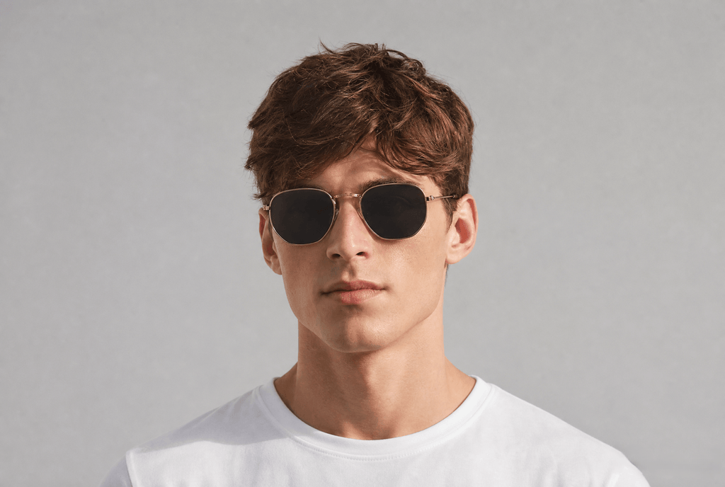 A man wearing round metal-framed sunglasses with dark lenses and a white t-shirt, photographed against a neutral light gray background.