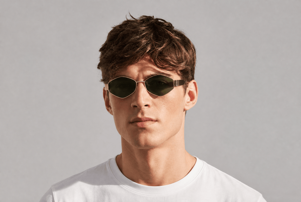 A man wearing oval dark sunglasses with a thin metallic frame and a white t-shirt, photographed against a neutral light gray background.  Gold/green