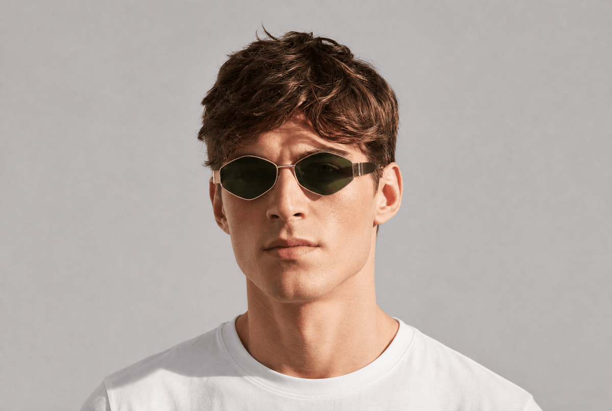 A man wearing oval dark sunglasses with a thin metallic frame and a white t-shirt, photographed against a neutral light gray background.  Gold/green