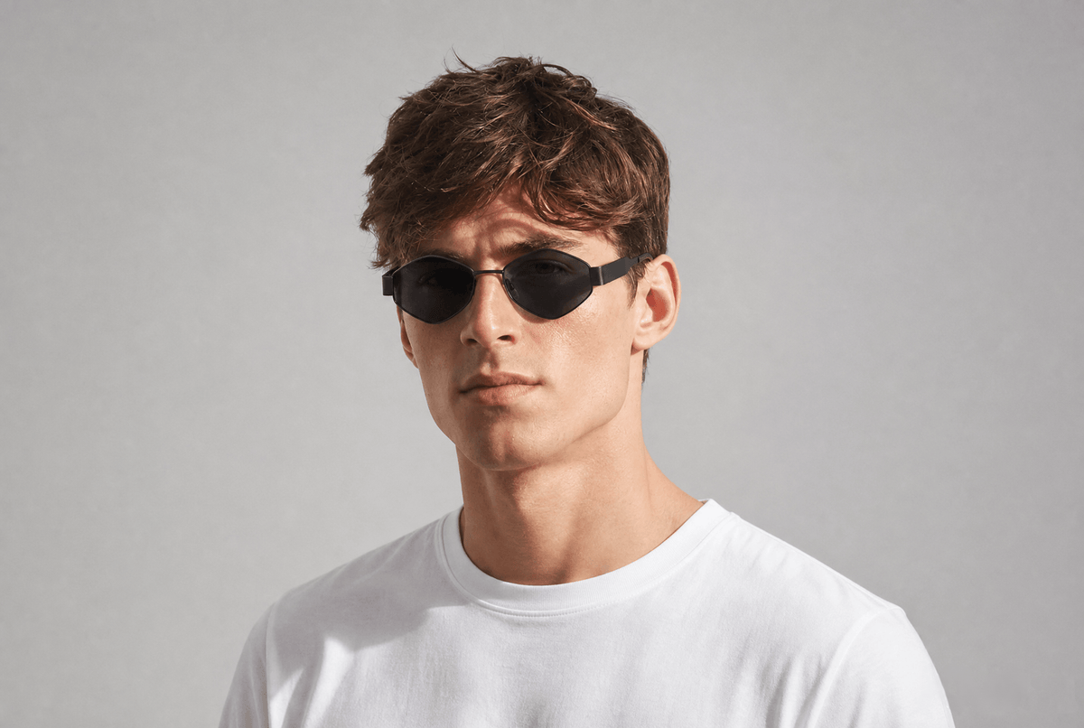 A man wearing black cat-eye sunglasses and a white t-shirt, photographed against a neutral light gray background. Black